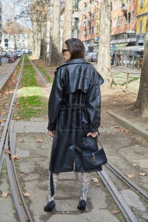 Photo for MILAN, ITALY - FEBRUARY 25, 2023: Woman with black leather trench coat with Missguided writing  and black Yves Saint Laurent bag before Dolce and Gabbana fashion show, Milan Fashion Week street style - Royalty Free Image