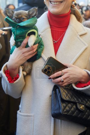Foto de MILÁN, ITALIA - 13 DE ENERO DE 2024: Mujer con bolso Chanel de cuero negro y perro de barbilla japonés con chaqueta hinchable verde antes del desfile de moda MSGM, Milan Fashion Week street style - Imagen libre de derechos