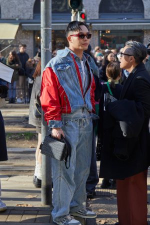 Téléchargez les photos : MILAN, ITALIE - 13 JANVIER 2024 : Homme avec pantalon en denim bleu et veste à manches rouges et sac à motifs logo avant le défilé Dolce et Gabbana, Milan Fashion Week street style - en image libre de droit