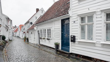 Foto de Stavanger, Noruega - 3 de agosto de 2018: el casco antiguo conocido como Gamble Stavanger con calles empedradas brillando bajo la lluvia se alineó con las tradicionales casas de madera blanca que datan del siglo XVIII - Imagen libre de derechos