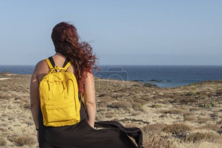 Rückansicht einer jungen hübschen Frau mit langen roten Haaren, einem schwarzen Kleid und einem leuchtend gelben Rucksack auf dem Rücken, während sie die Küstenlandschaft von Abades auf Teneriffa, Kanarische Inseln, Spanien betrachtet