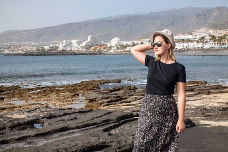 Foto de Hermosa mujer caucásica con gafas de sol y un sombrero mientras disfruta de un paseo por la costa volcánica de Las Américas, con la vista del complejo turístico en la parte posterior, Tenerife, Islas Canarias, España - Imagen libre de derechos