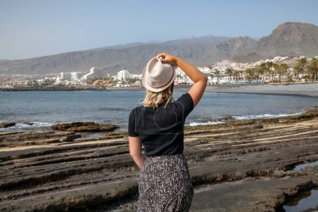 Foto de Vista trasera de la joven mujer caucásica con colores oscuros y sosteniendo su sombrero con una mano mientras mira hacia la costa de Las Américas, destino turístico favorito, Tenerife, Islas Canarias, España - Imagen libre de derechos