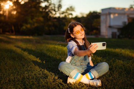 Téléchargez les photos : Enfant fille prendre un selfie par téléphone mobile en plein air dans le parc naturel vert - en image libre de droit