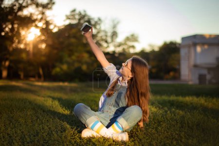 Téléchargez les photos : Adorable enfant fille faire selfie sur téléphone portable assis dans l'herbe profonde dans le parc - en image libre de droit