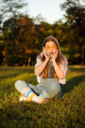 Foto de Sorprendida y emocionada chica de la escuela en overoles de mezclilla hablar en el teléfono inteligente sentado en la hierba verde en el parque - Imagen libre de derechos