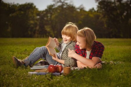 Foto de Dos hermanos leen libro juntos sobre hierba verde en el parque al aire libre en el día soleado - Imagen libre de derechos