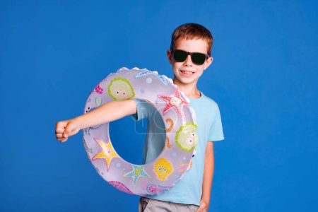 Téléchargez les photos : Garçon dans les lunettes de soleil tenant anneau gonflable prêt ti visite piscine sur fond bleu clair studio - en image libre de droit