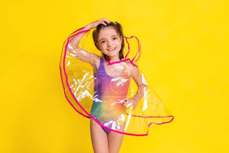 Téléchargez les photos : Enfant fille portant en imperméable transparent et en maillot de bain arc-en-ciel sur fond jaune, prêt pour les douches tropicales pluvieuses - en image libre de droit
