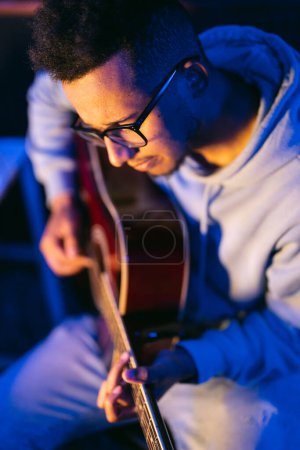 Téléchargez les photos : Jeune musicien afro portant des lunettes jouant de la guitare acoustique, sur fond sombre - en image libre de droit