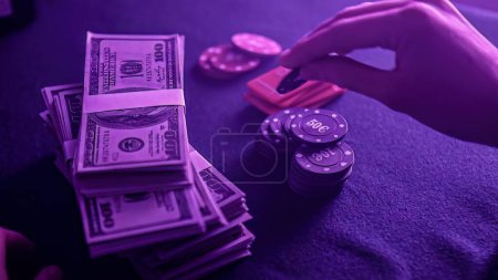 Téléchargez les photos : Stack of poker chips in a range of colors and a wad of dollars on the gaming table in a dimly lit casino. The chips and money suggest a game of poker is about to begin. - en image libre de droit