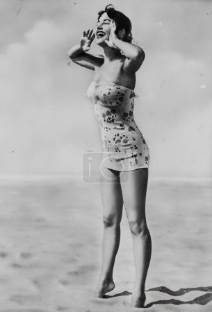 Foto de Roma, Italia mayo 1951: Una joven impresionante hace alarde de sus curvas en un mini vestido de inspiración vintage mientras descansa en una playa de arena. - Imagen libre de derechos