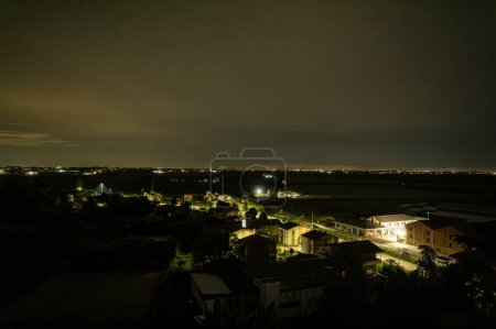Foto de Encantadora vista aérea nocturna de un pequeño pueblo italiano enclavado en el sereno valle del Po, brillando bajo el cielo iluminado por la luna. - Imagen libre de derechos