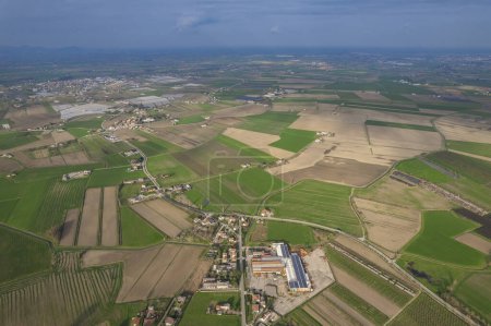 The aerial view showcases vast farmland in the Po Valley region. Fields are neatly divided into plots with crops growing, visible farmhouses, barns, and irrigation systems.