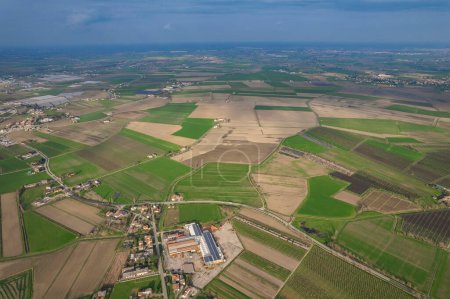 This aerial view shows a vast expanse of farmland in Pianura Padana, with neatly planted crops and irrigation systems. In the background, a cityscape can be seen with tall buildings and bustling streets.