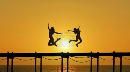Téléchargez les photos : Deux femelles silhouettées bondissent dans les airs, leurs figures dessinées contre un ciel magnifique au coucher du soleil, alors qu'elles sautent sur une jetée - en image libre de droit