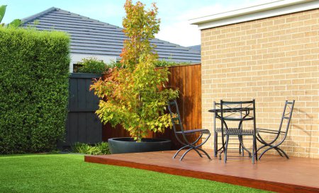 Australian backyard showcases a lush artificial grass lawn and a majestic tree in the background, providing a natural touch against a wooden fence, wooden decking area furnished with stylish outdoor furniture, all set against brick wall