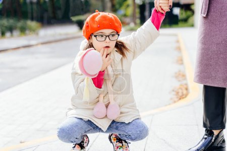 Photo for Cute girl in coat red beret in eyeglasses with special needs holding toy and mothers hand on a walk outdoor. - Royalty Free Image