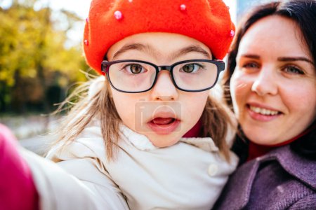 Photo for Cute curious little daughter in eyeglasses wearing red beret and mother are doing selfie outdoor in the city street. Happy family moments concept. - Royalty Free Image
