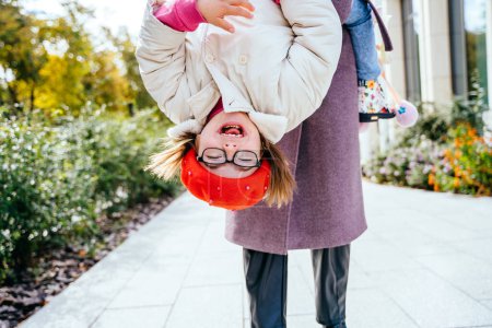 Photo for Brunette mom and cheerful adorable tot girl playing, having fun together in city street in autumntime, mother playfully holding her cute little daughter upside down. - Royalty Free Image