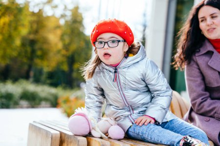 Photo for Happy family moments in the city. Cute curious little daughter in eyeglasses wearing red beret and mother are sitting at bench outdoor in the city street. - Royalty Free Image