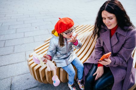 Photo for Brunette caucasian positive woman and daughter using smartphone sitting on the bench at city street. Mother calms the girl showing cartoon on the phone. Happy family moments in the city. - Royalty Free Image
