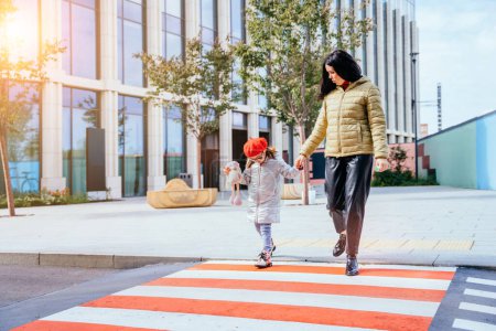 Photo for Young caucasian mother crossing street and commuting downtown city with little daughter wearing eyeglasses and red beret in autumn time. Full height. - Royalty Free Image