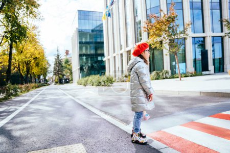 Téléchargez les photos : Jolie petite fille avec un jouet vêtu d'un manteau et béret rouge se tient seul près du passage supérieur à la rue de la ville. - en image libre de droit