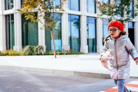 Téléchargez les photos : Jolie petite fille handicapée avec un jouet portant un manteau et un béret rouge courir tout en traversant la route à travers le passage pour piétons. Copier le texte espace - en image libre de droit