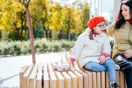 Photo for Portrait of young mother who sitting on bench outdoor with cute beautiful disabled daughter and smiling, communication to each other. - Royalty Free Image