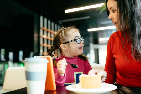 Photo for Portrait of happy mother smiling to each other cute girl with down syndrome eating cake in cafe. Happy family moments concept. - Royalty Free Image