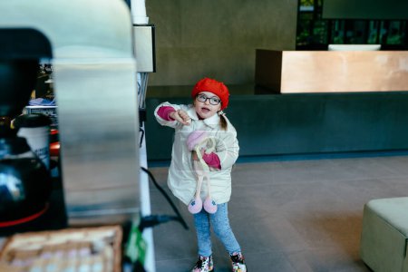 Photo for Cute little girl in eyeglasses red beret and coat indoor in lobby or coffee shop. Children hold a toy. - Royalty Free Image
