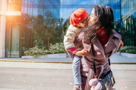 Photo for Cute young daughter on a piggy back ride with her mother in autumn time with modern building at city street on background. Sun glare efect - Royalty Free Image