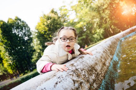 Foto de Retrato de una hermosa joven con dos trenzas en gafas, abrigo blanco mirando a un lado sonriente disfrutando del buen tiempo soleado en el parque de otoño al aire libre. Efecto de deslumbramiento solar - Imagen libre de derechos