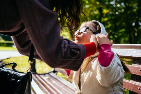 Téléchargez les photos : Mère méconnaissable mettant écouteurs sa petite fille handicapée dans des lunettes pour se calmer en plein air dans le parc. Syndrome de Down dans la vie quotidienne. - en image libre de droit