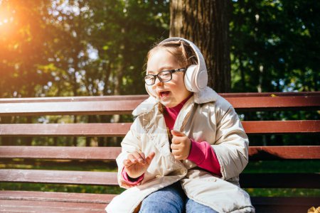 Téléchargez les photos : Portrait de jolie petite fille mignonne écoutant de la musique avec des écouteurs assis sur le banc au parc en plein air. Effet éblouissement solaire. - en image libre de droit