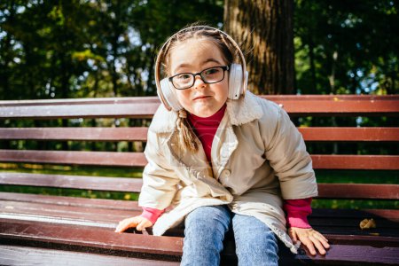 Photo for Beautiful little girl in eyeglasses with down syndrome smiling and looking into camera with cute facial expression. Child with headphones sitting on bench at sunny autumn park outdoor. - Royalty Free Image