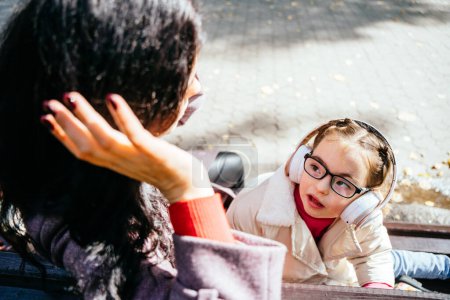 Photo for Life in the education age of special children, Happy disability kid concept. Cute little girl in earphones looking at her mother relax on bench at park outdoor. - Royalty Free Image
