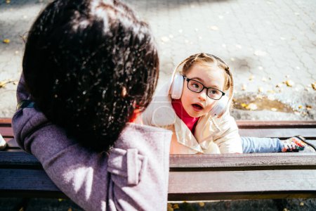 Téléchargez les photos : Jolie petite fille mignonne avec handicap écoutant de la musique avec expression de rêve sur le visage et la mère dans le parc en plein air. - en image libre de droit
