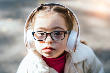 Photo for Close up portrait of beautiful little girl in eyeglasses with down syndrome smiling and looking into camera with thoughtful facial expression. Child with headphones at sunny autumn park outdoor. - Royalty Free Image