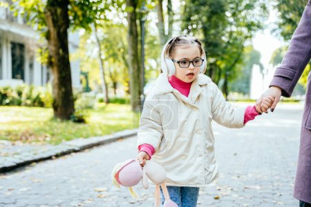 Téléchargez les photos : Jolie fille avec des besoins spéciaux dans les lunettes portant un manteau, écouteurs tenant les mères main sur une promenade en plein air. - en image libre de droit