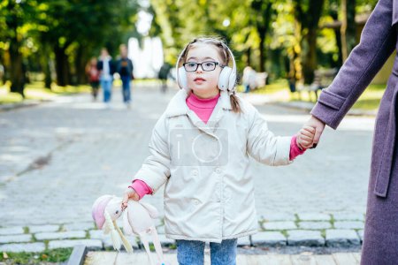 Téléchargez les photos : Besoin spécial enfant tenant les mains des mères et jouet portant des écouteurs s'amuser dans le parc de la ville en automne. - en image libre de droit