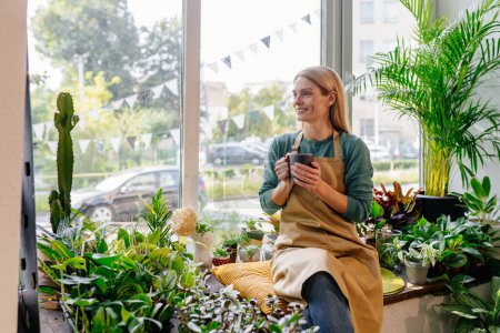 Photo for Business female owner relaxing drinking coffee at flower shop sitting surrounded by plants at windowsill looks positive, not afraid of challenges. The concept of starting a business. - Royalty Free Image