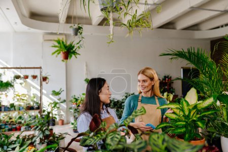 Foto de Dos mujeres de diferentes edades y nacionalidades están conversando mientras trabajan, cuidando plantas en una tienda llena de flores de colores brillantes. - Imagen libre de derechos