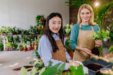Photo for Portrait of business owner and her assistant wear uniforms, different ages and nationalities, are conversing while working together, caring for plants in a brightly colored flower-filled store. - Royalty Free Image