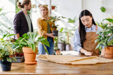 Photo for Charming Asian female worker in apron working behind counter, couple choosing plant in pot on blurred background. Plants business, start up. Small business concept. - Royalty Free Image