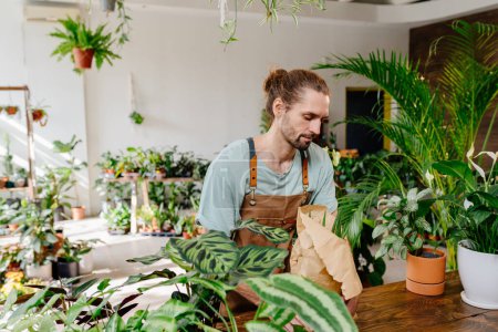 Photo for Handsome Caucasian man wearing apron wrapping a potted plant in paper with care in greenery at floral shop. Enjoying his job at cozy flower shop. Urban jungle and plant parent concept. - Royalty Free Image