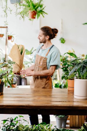 Foto de Vendedor caucásico tiro vertical, empleado con delantal venta de plantas en maceta en una tienda de flores. - Imagen libre de derechos