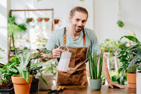 Photo for Handsome young man wearing apron carefully watering potted houseplants indoors. Concept of plants care and home garden. Small business entrepreneur. Urban jungle and plant parent concept. - Royalty Free Image