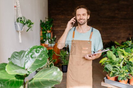 Photo for Worker in uniform carrying flower in pot for customer and talking on mobile phone while working in flower shop - Royalty Free Image